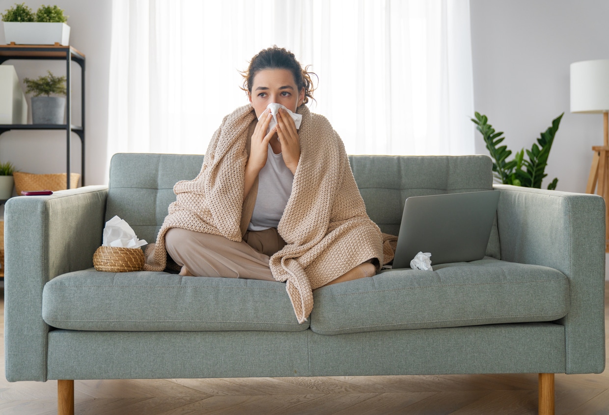 A girl is holding a paper tissue and blowing her nose while sitting on a gray couch.