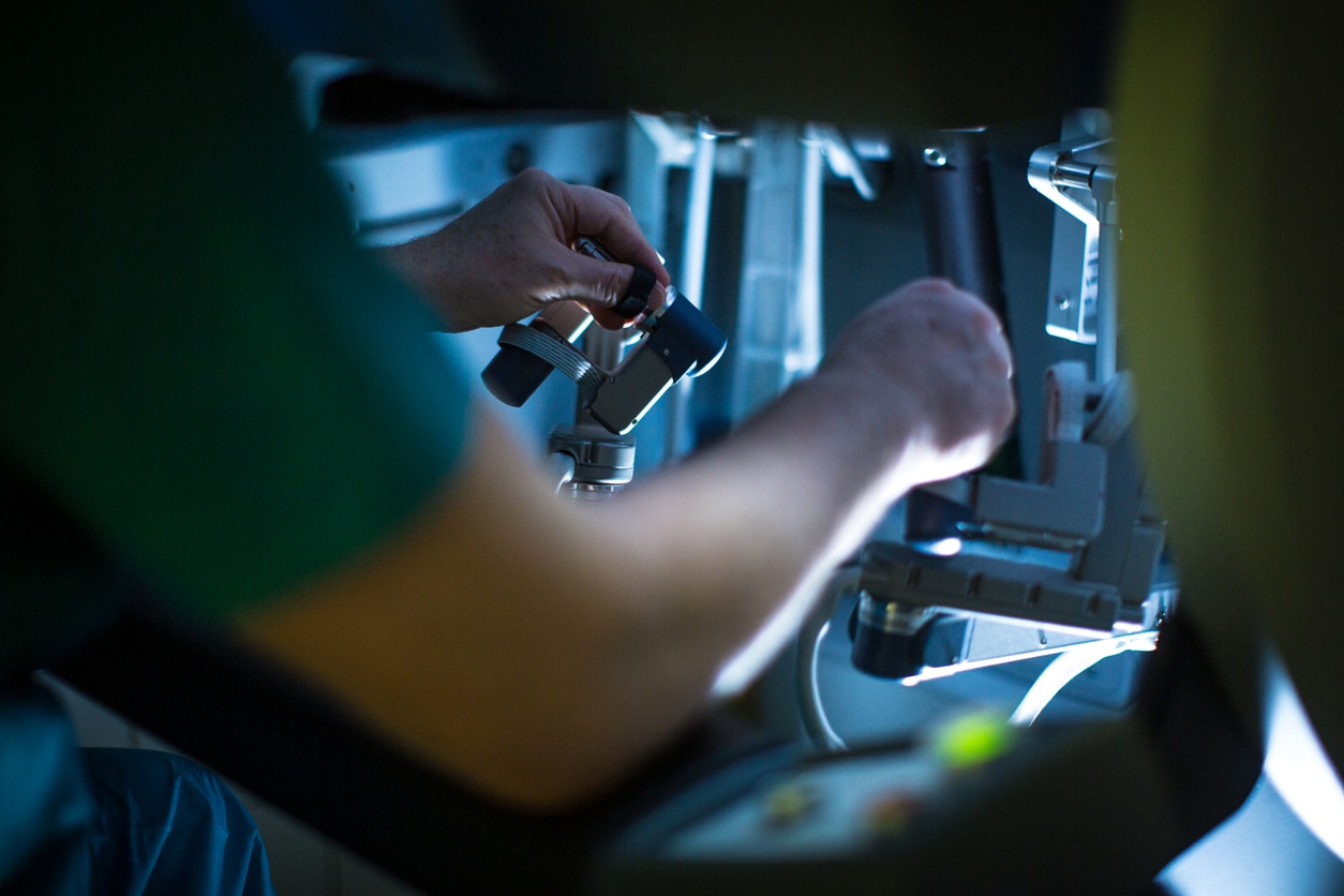 A surgeon in blue scrubs operating a robotic surgery system with two hands.