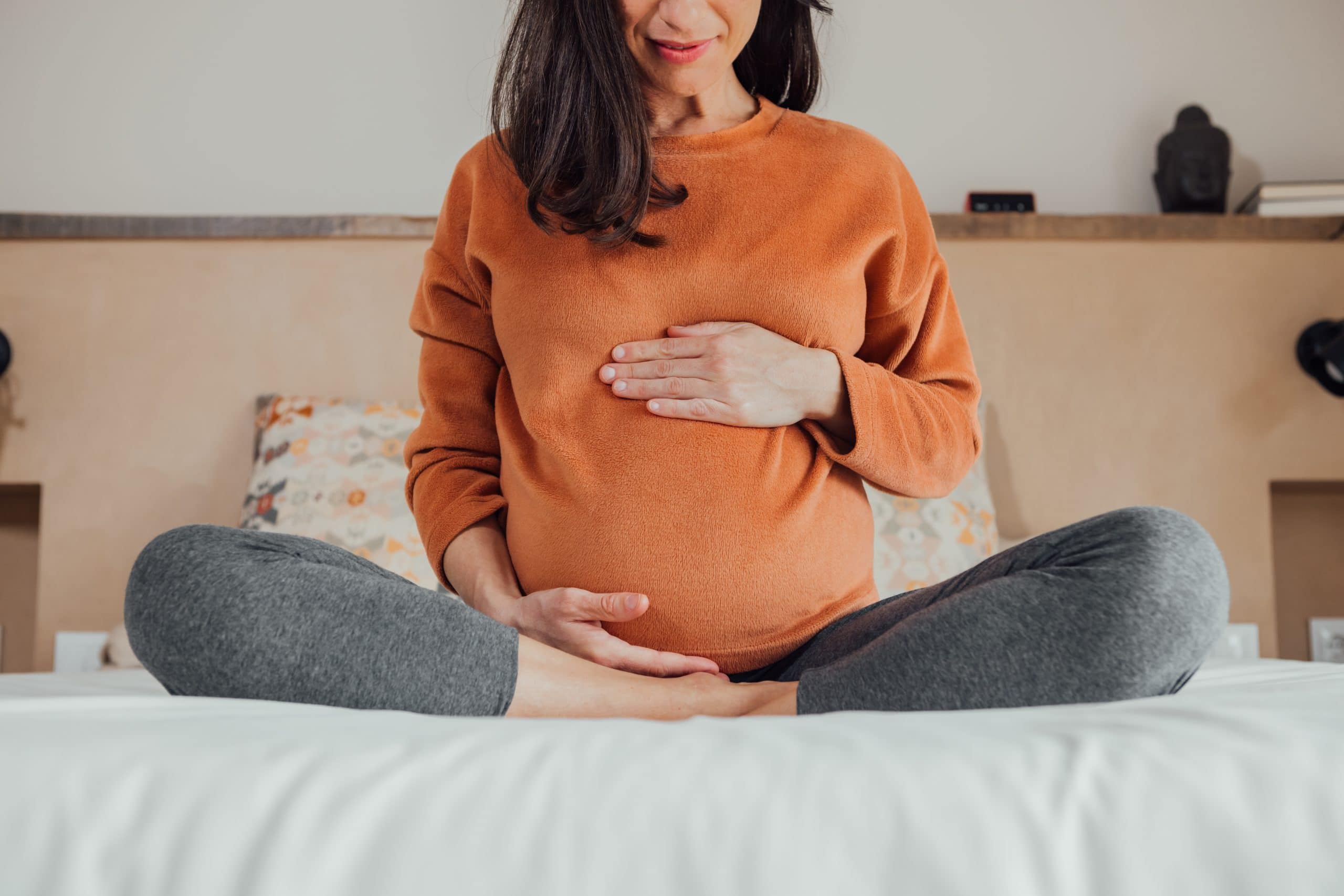 A pregnant woman sits on a bed with legs crossed holding her belly, representing endometriosis and pregnancy