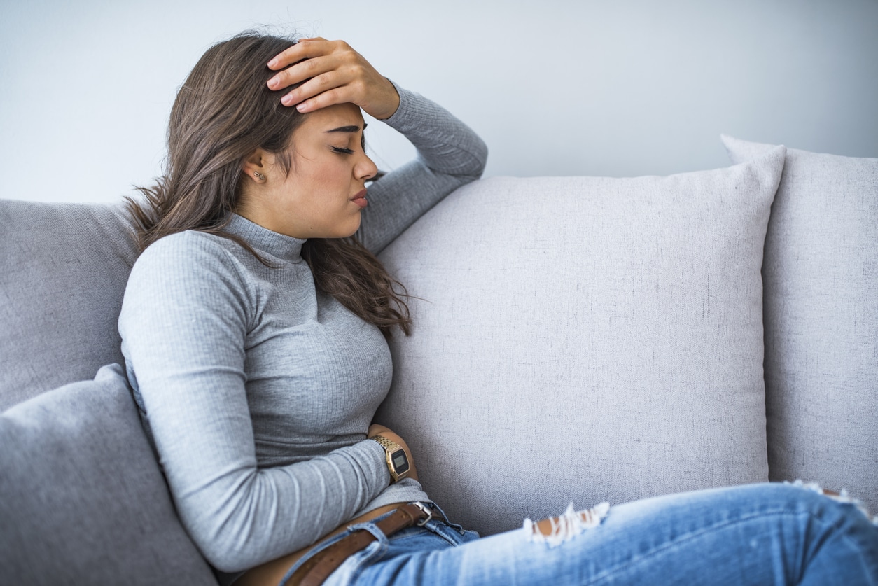 A brunette woman in a gray shirt and jeans sitting on the couch with her hand on her forehead and stomach.