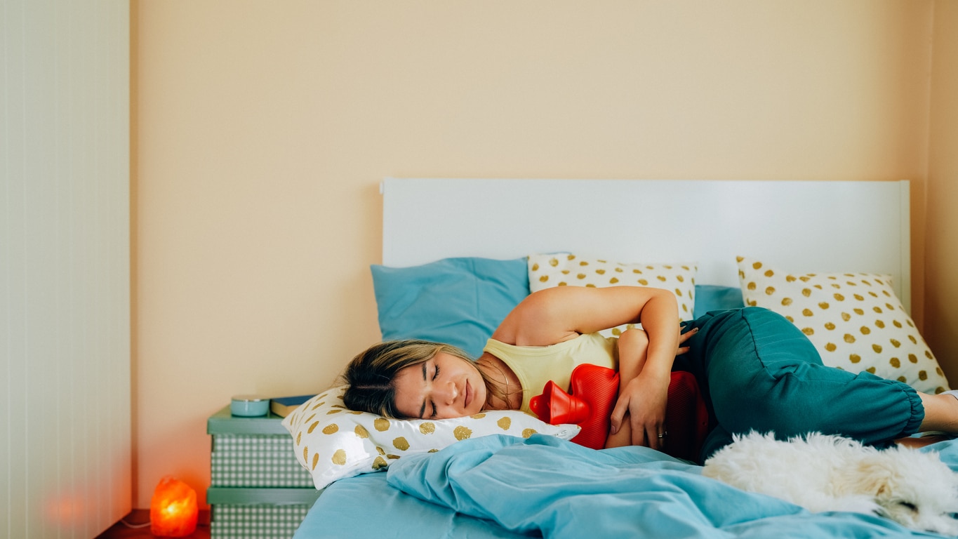 A young woman lying on a blue and yellow bed with a heating pad over her abdomen.