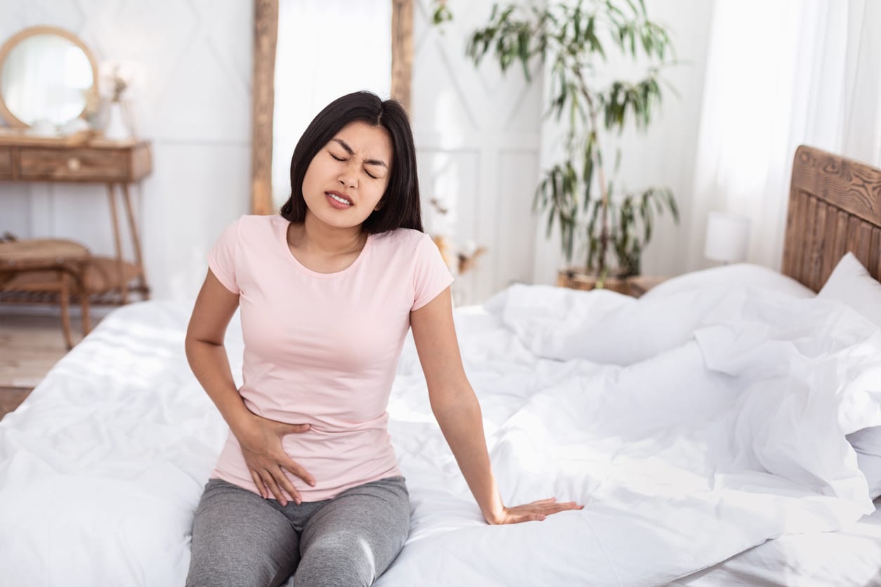 A woman in a light pink shirt and gray pants sitting on the bed with her hand on her abdomen due to pelvic pain.