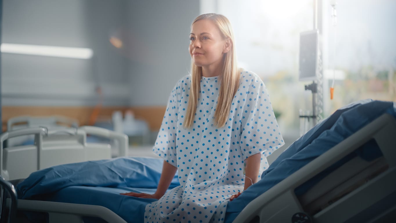 A blonde girl in a hospital gown sitting on the edge of her bed after a full recovery.