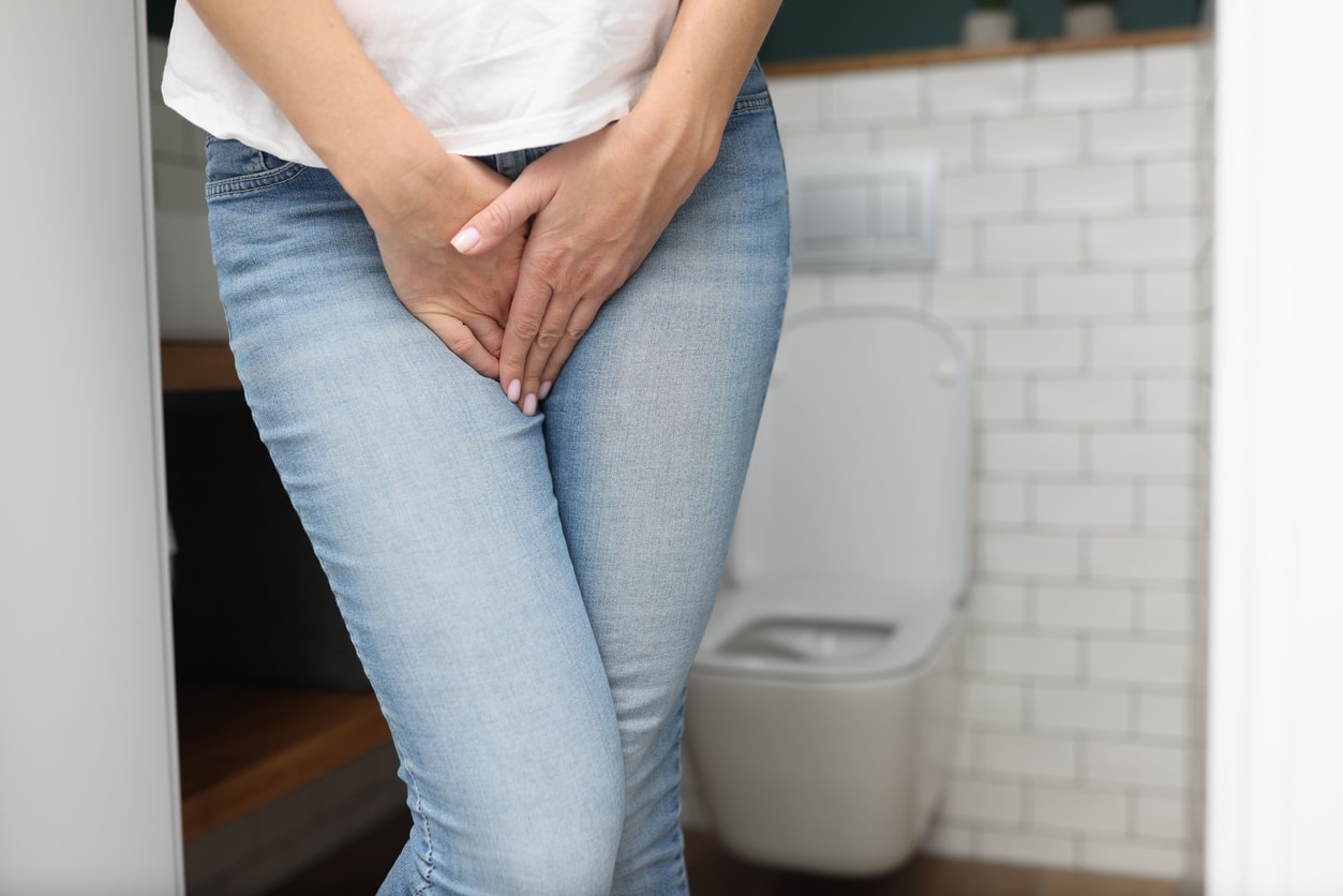 Female in a white shirt with her hands over her bladder in blue jeans.