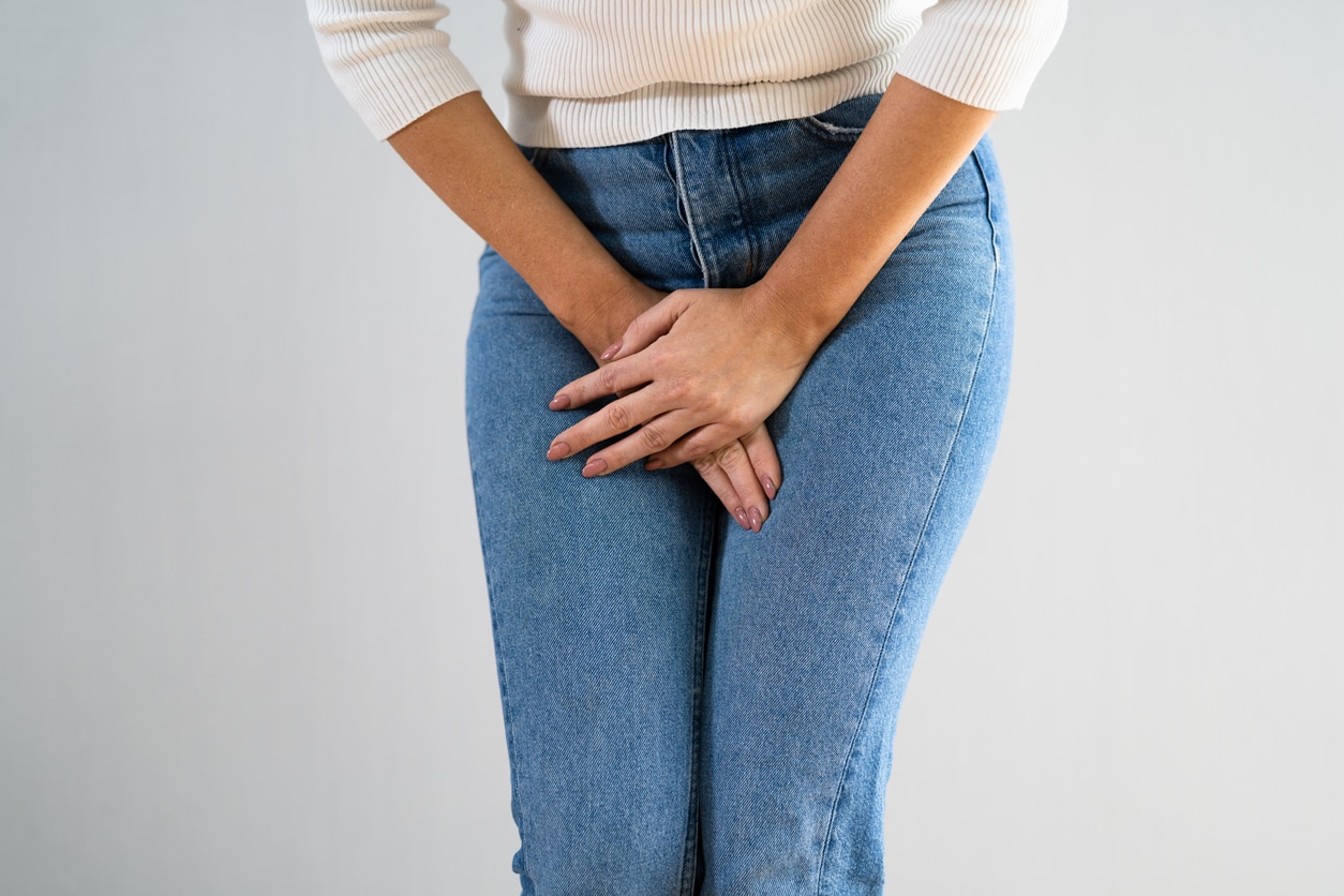 A woman in a white sweater and blue jeans holding her bladder on a white background.